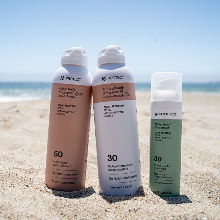 Three sunscreen bottles on a sandy beach with a clear sky.
